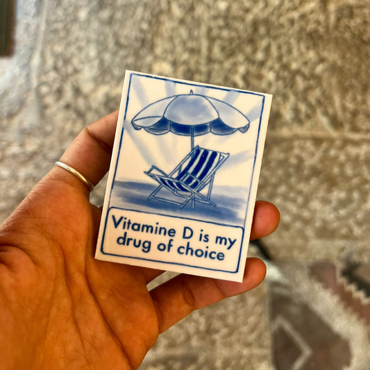 Hand holding a small card with a sunbed and umbrella illustration and text 'Vitamine D is my drug of choice' against a textured background.