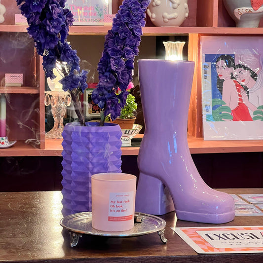 Purple glossy rain boot with a candle on a wooden surface, with decorative elements and posters in the background.