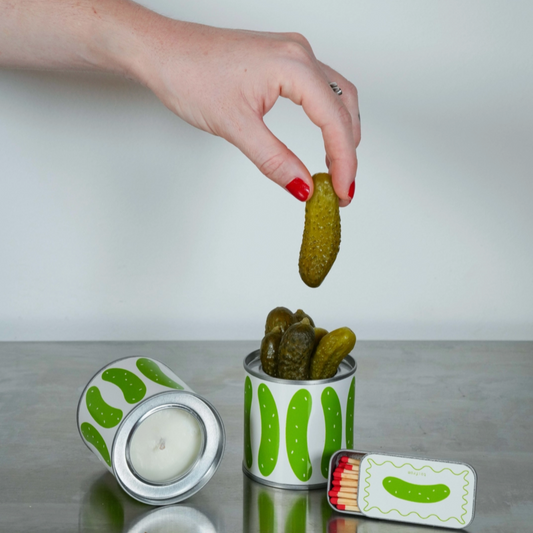 Hand holding a pickle over a pickle-shaped candle with a matchbox featuring a pickle design on a gray surface.