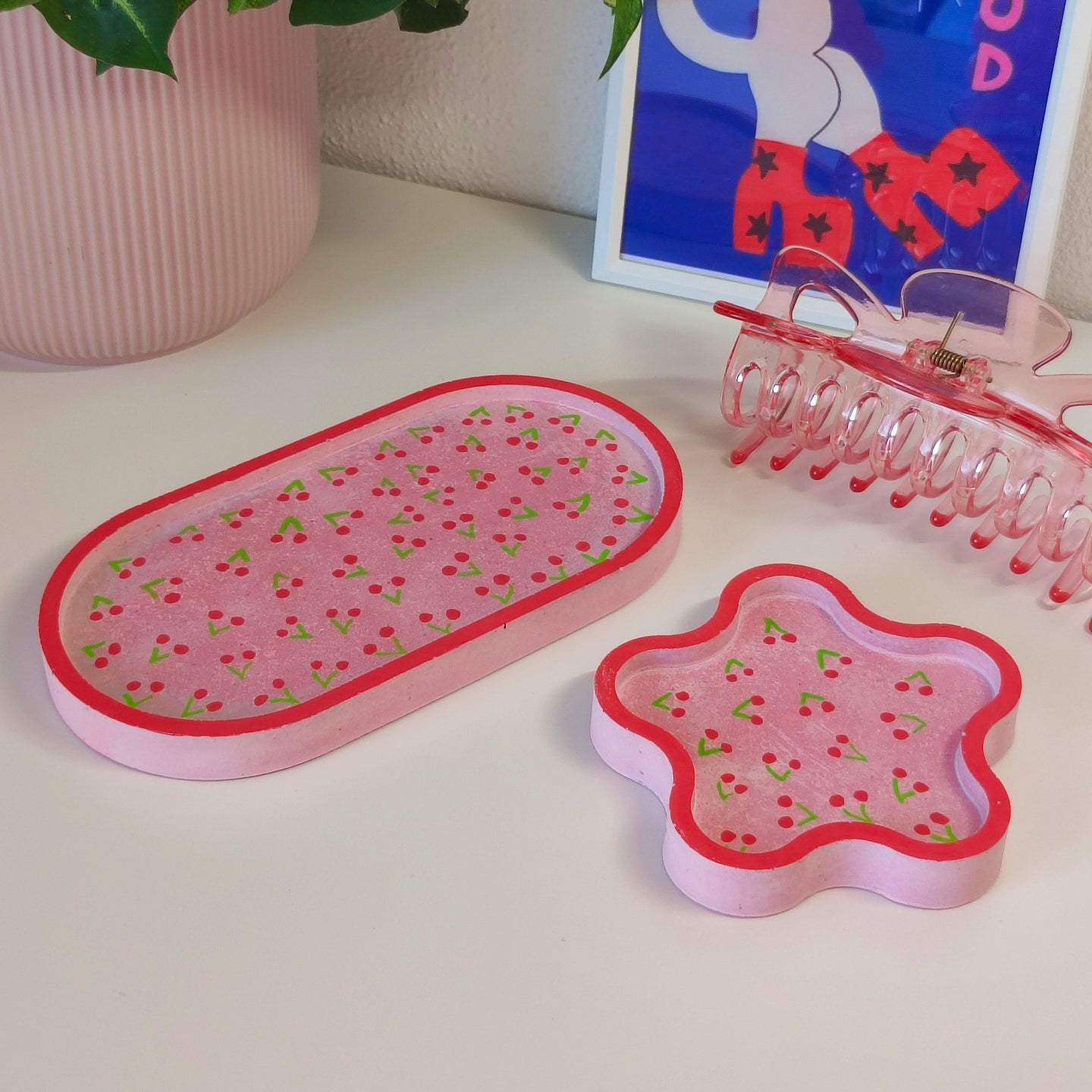 Pink hair accessories including a clip and a comb on a white surface with a plant and framed picture in the background.