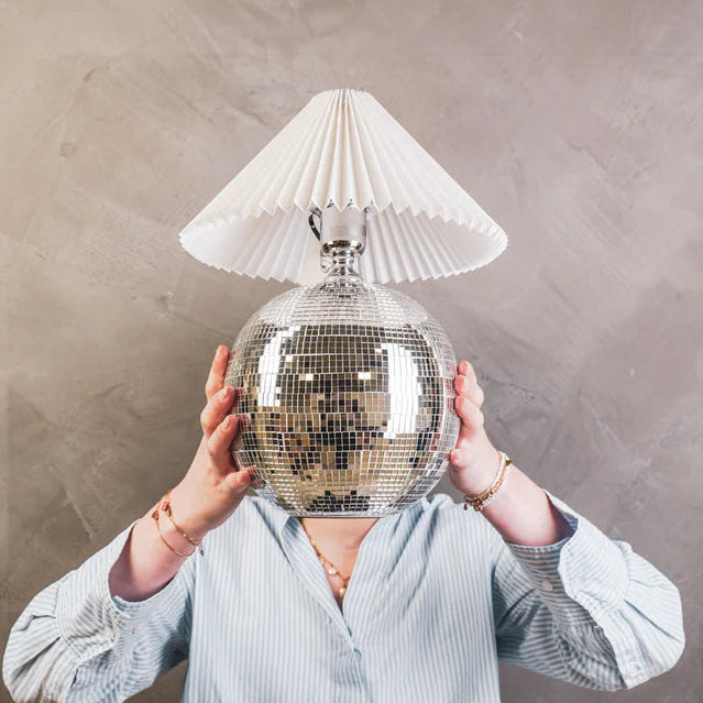 Person holding a disco ball with a lampshade over their head against a plain background