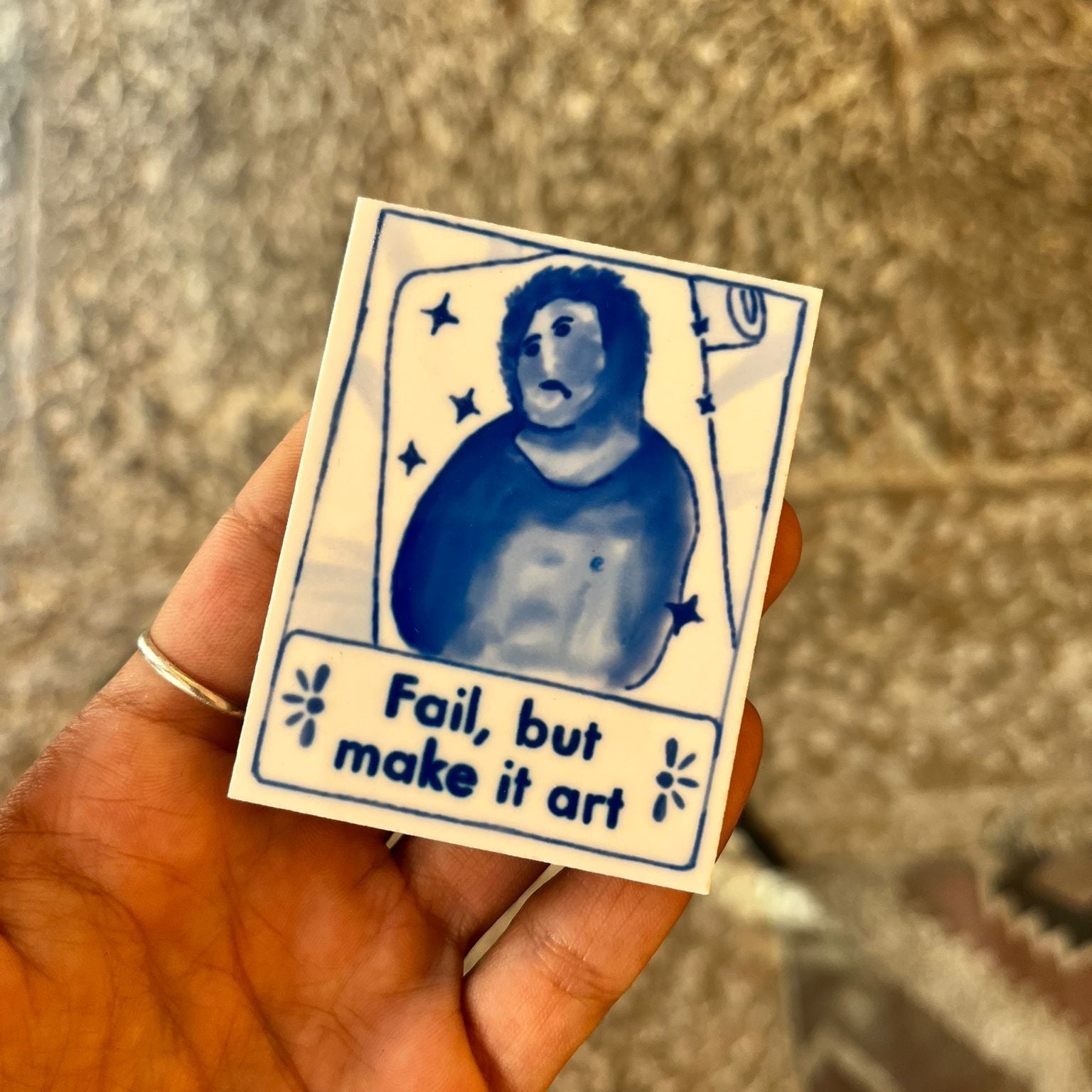 Hand holding a small card with a blue illustration of a person and text 'Fail, but make it art' on a textured surface.