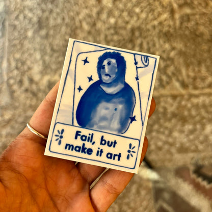 Hand holding a small card with a blue illustration of a person and text 'Fail, but make it art' on a textured surface.