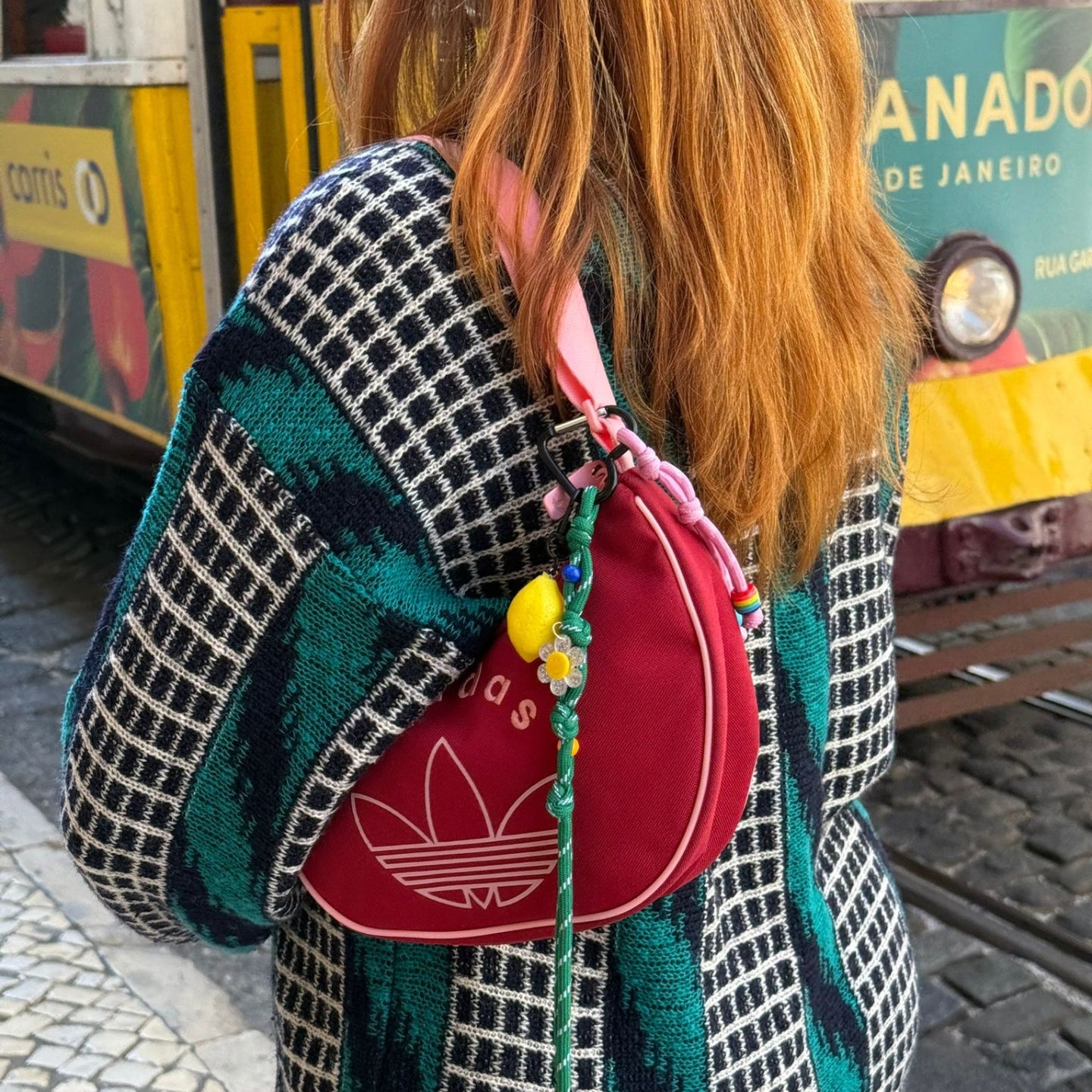 Person with a colorful patterned coat and red Adidas bag walking past a tram in an urban setting.