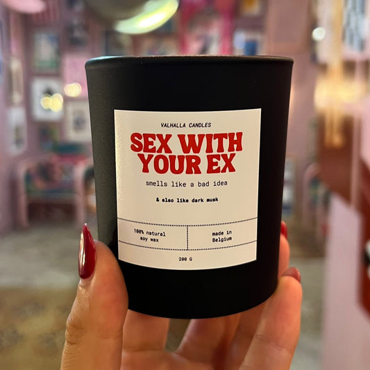 Black candle with a label reading 'Sex with Your Ex' held in front of a blurred indoor background.