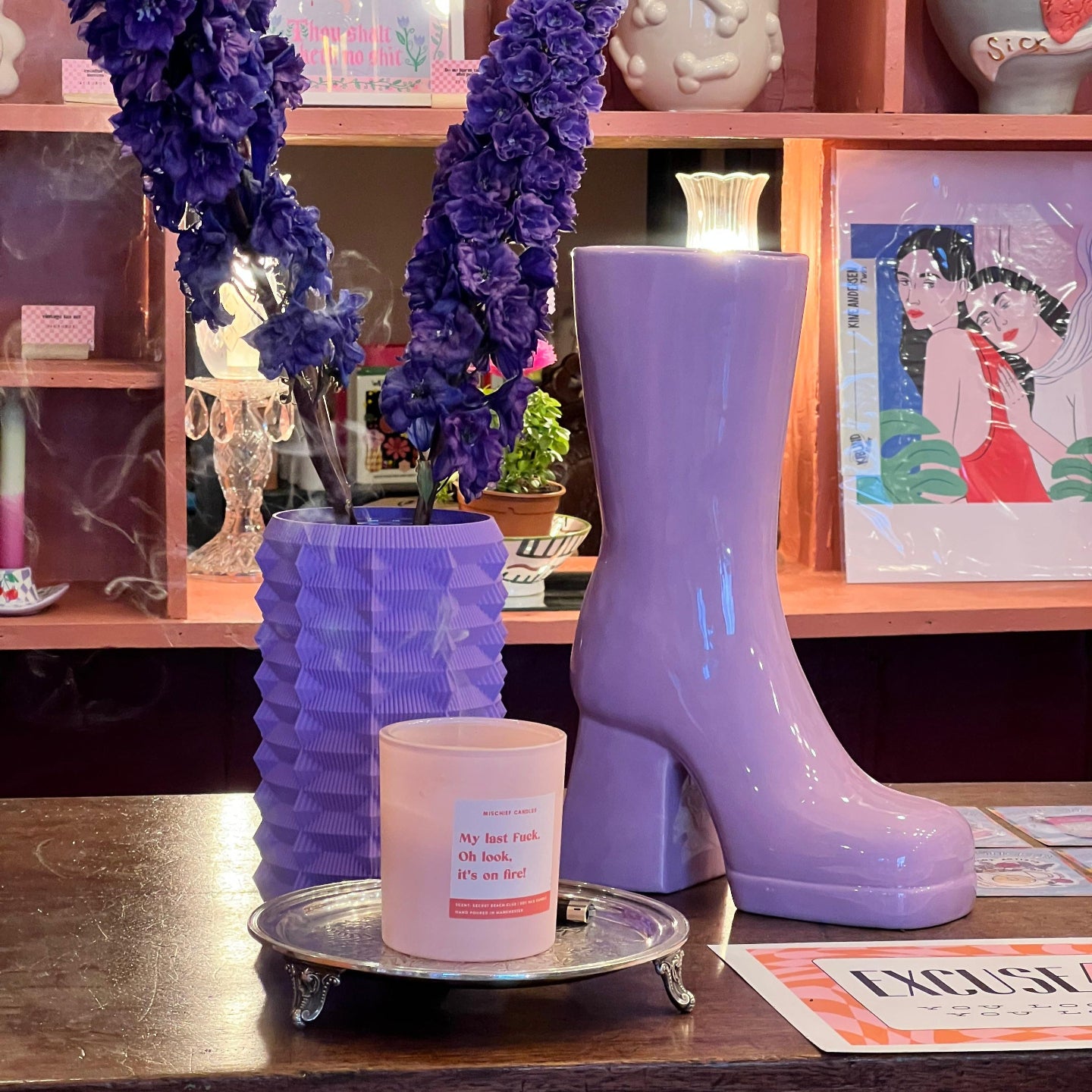 Purple glossy rain boot with a candle on a wooden surface, with decorative elements and posters in the background.