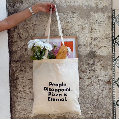 Tote bag with 'People Disappoint. Pizza is Eternal.' text held by a person against a textured wall.