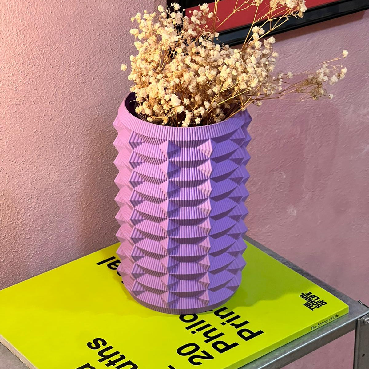 Purple textured vase with dried flowers on a green book against a pink wall