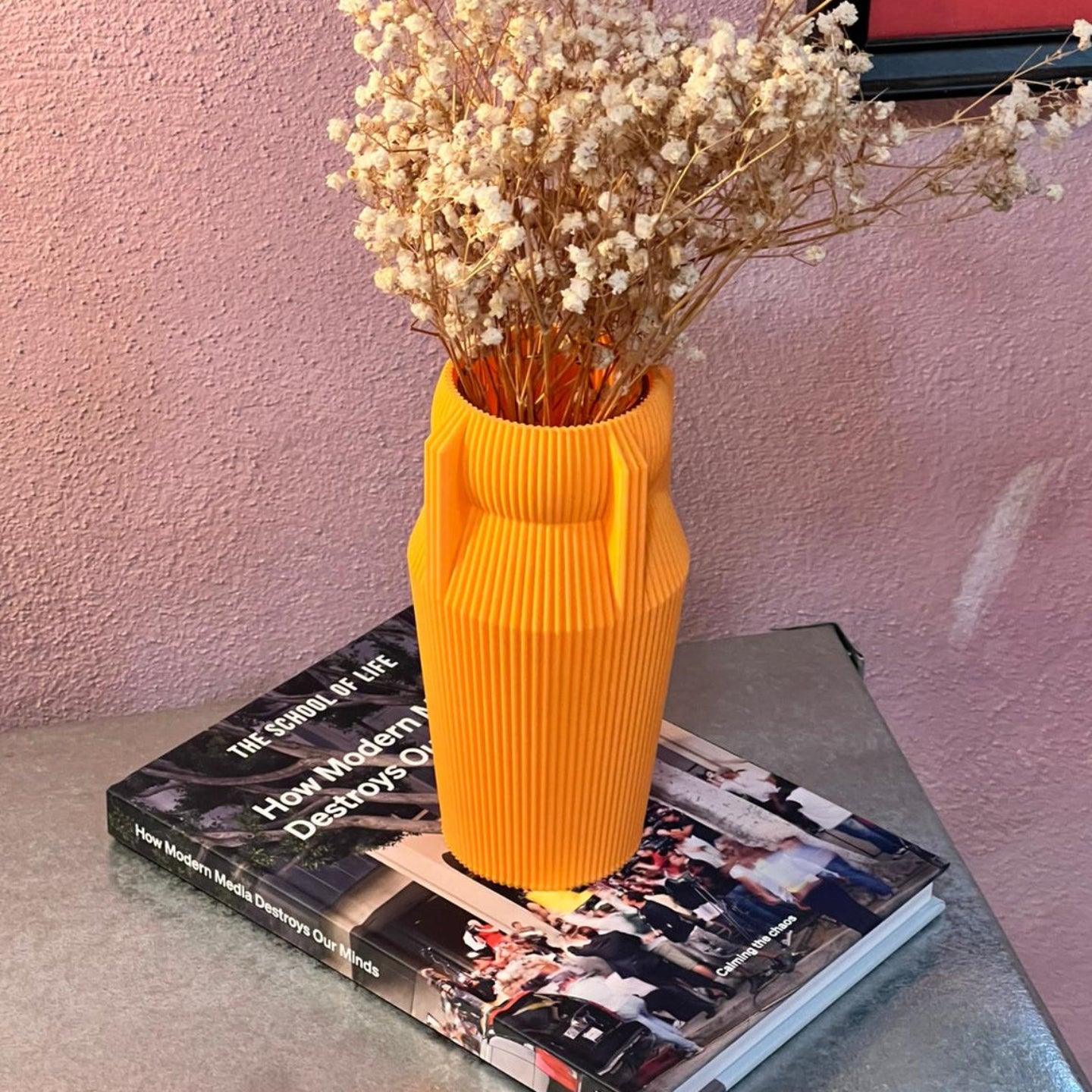 Yellow vase with dried flowers on a book against a pink wall