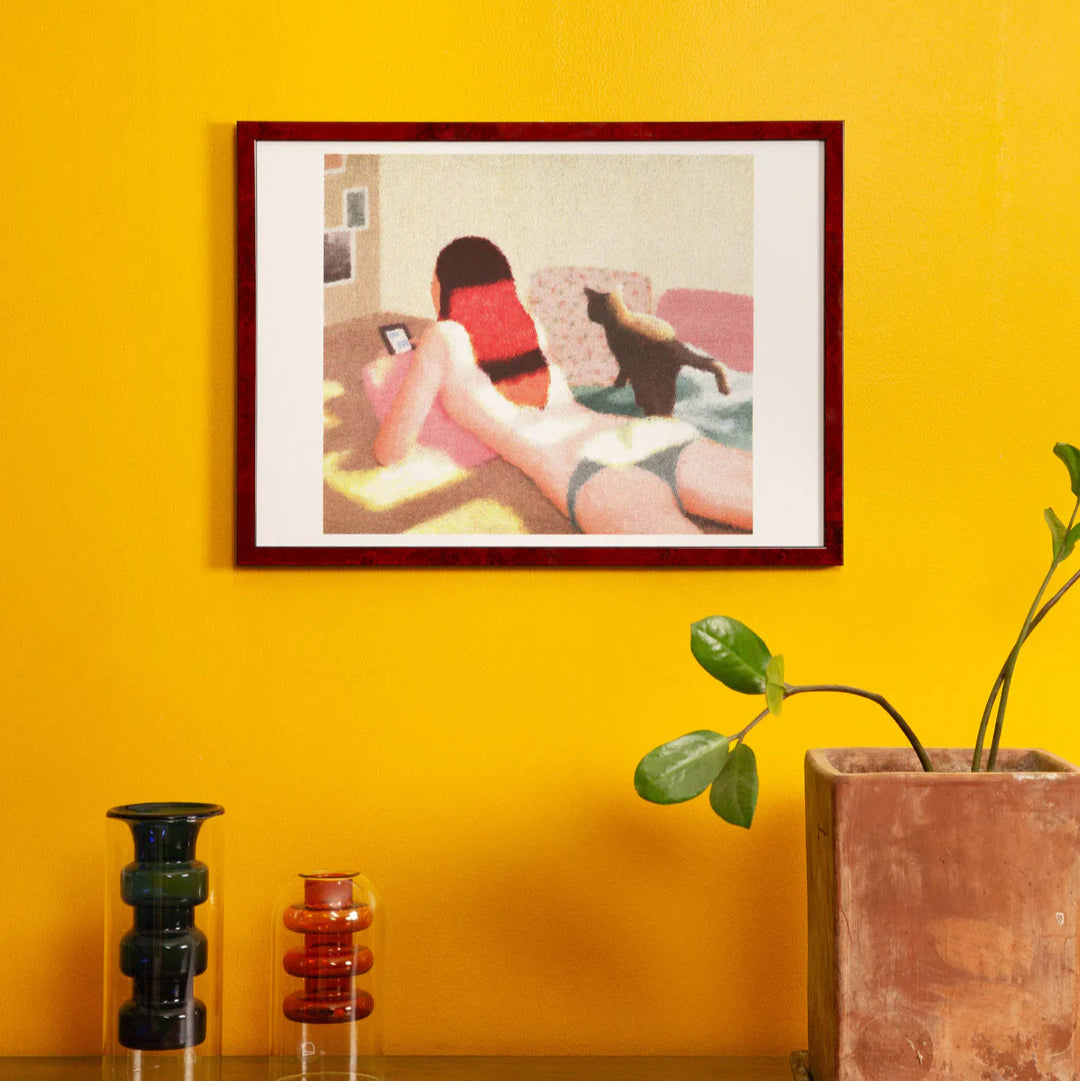 Framed artwork of a woman on a couch against a yellow wall with decorative vases and a plant.