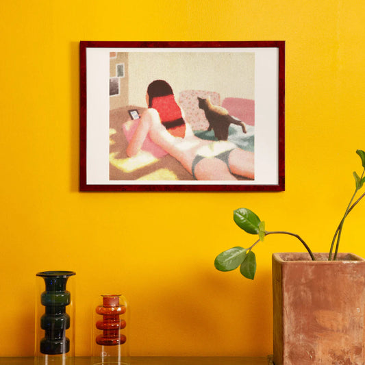 Framed artwork of a woman on a couch against a yellow wall with decorative vases and a plant.