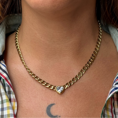 Close-up of a person wearing a gold chain necklace with a heart pendant, gold hoop earrings, and a colorful plaid shirt.