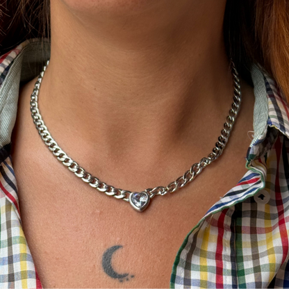 Person wearing a silver chain necklace with a heart pendant, visible tattoo on shoulder, and plaid shirt.