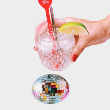 Hand holding a glass with ice and a lime wedge, featuring a heart-shaped gin stirrer against a pink background.